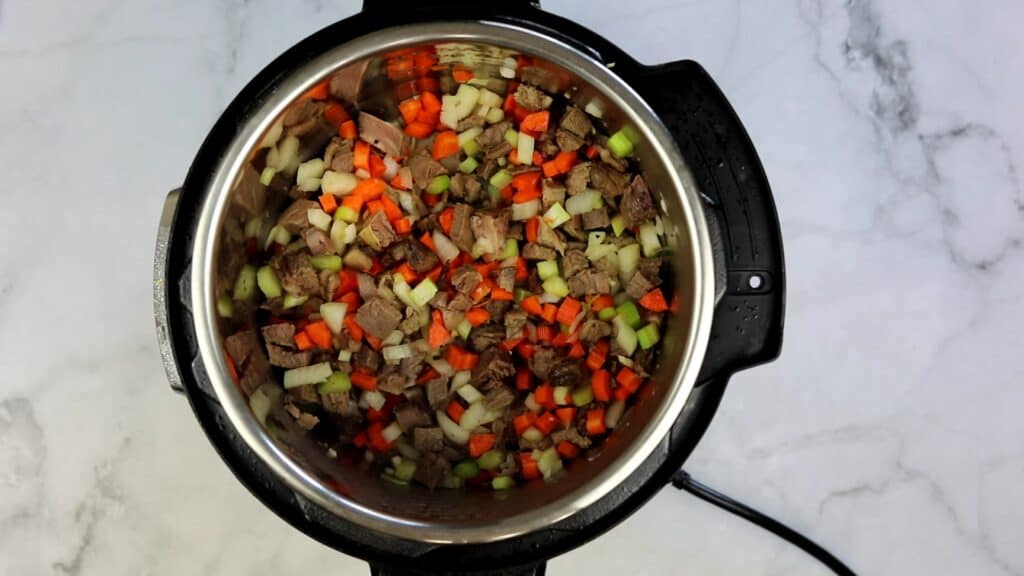 Chopped beef, carrots, celery, and onions are cooking in an Instant Pot on a marble countertop, coming together for a cozy homemade beef & barley soup.
