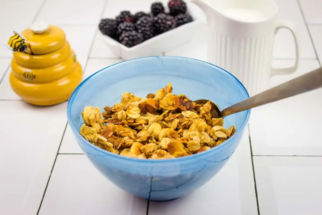 honey almond granola in a blue bowl with a spoon and berries in the background