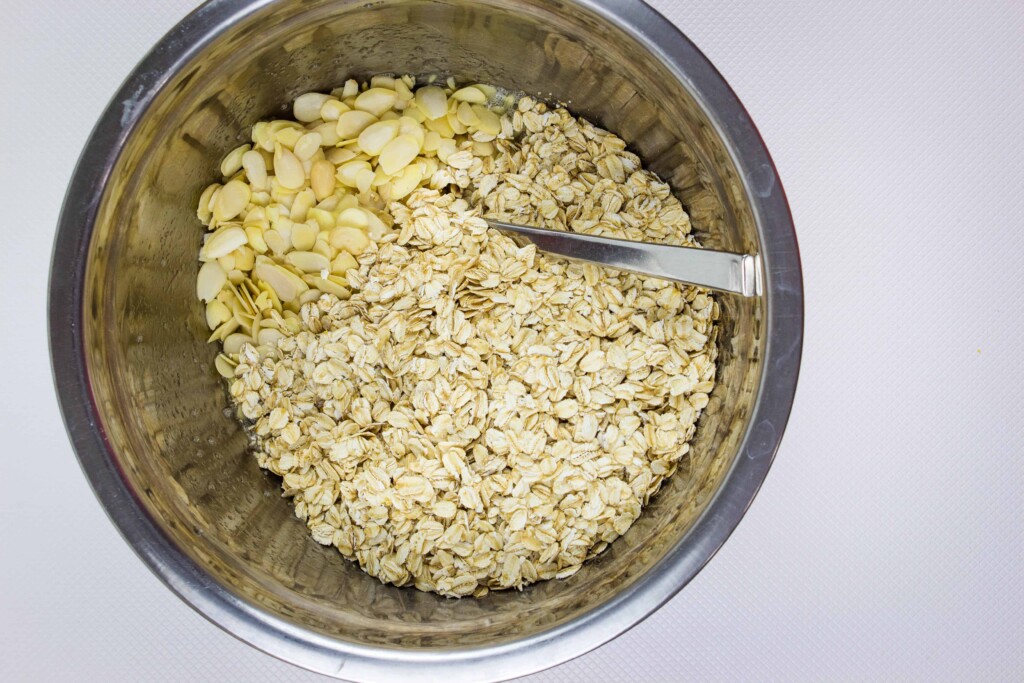 A metal bowl containing honey almond granola with rolled oats and sliced almonds, with a metal spoon resting inside.
