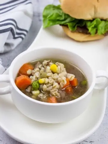 a bowlful of homemade beef & barley soup in a white bowl on a plate with a sandwich in the background