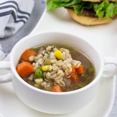 a bowlful of homemade beef & barley soup in a white bowl on a plate with a sandwich in the background