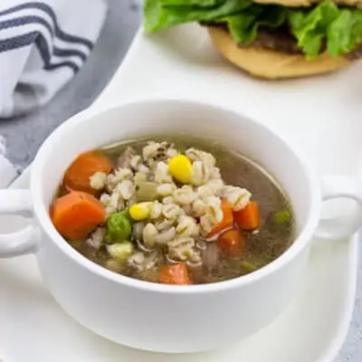 a bowlful of homemade beef & barley soup in a white bowl on a plate with a sandwich in the background