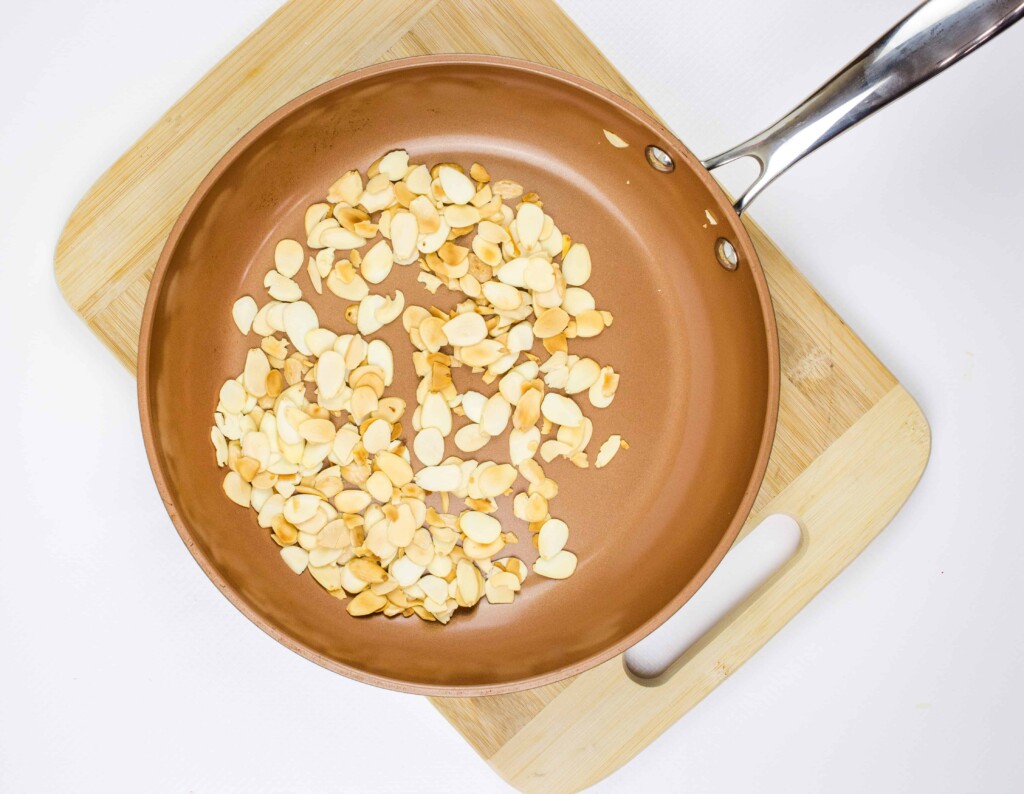 A frying pan with toasted almond slices, perfect for topping brussels sprouts with almonds, is placed on a wooden cutting board.
