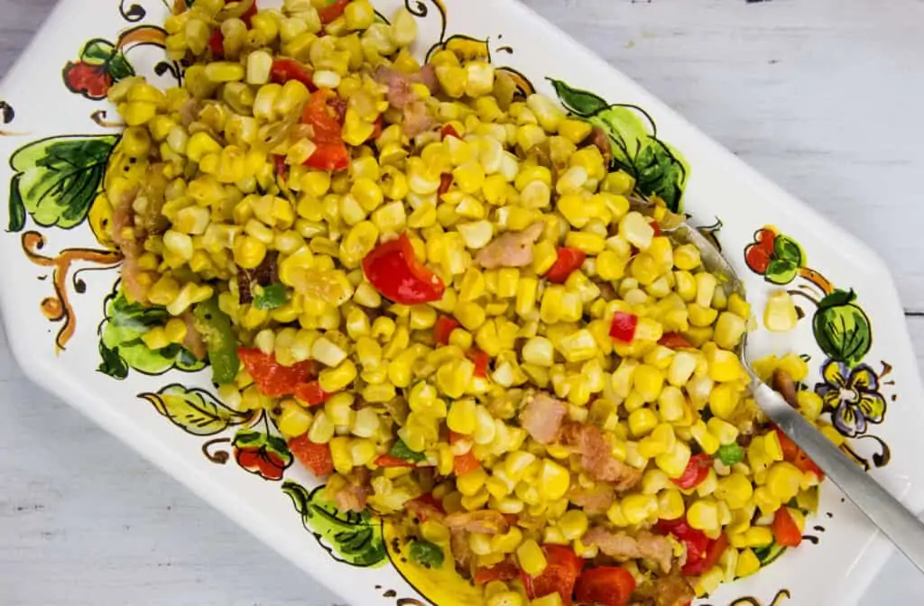 A rectangular dish filled with a corn salad mixed with red bell peppers and small pieces of bacon, with a serving spoon on the side.