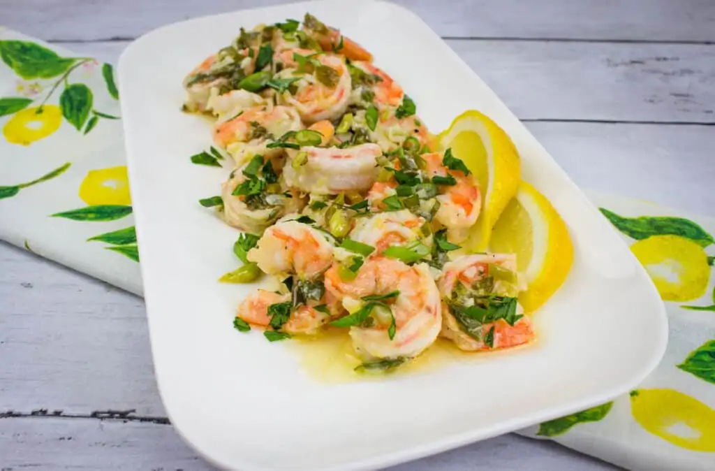 Shrimp sautéed with herbs and vegetables in a garlic shrimp scampi style, garnished with chopped parsley and served with lemon slices on a rectangular white plate.