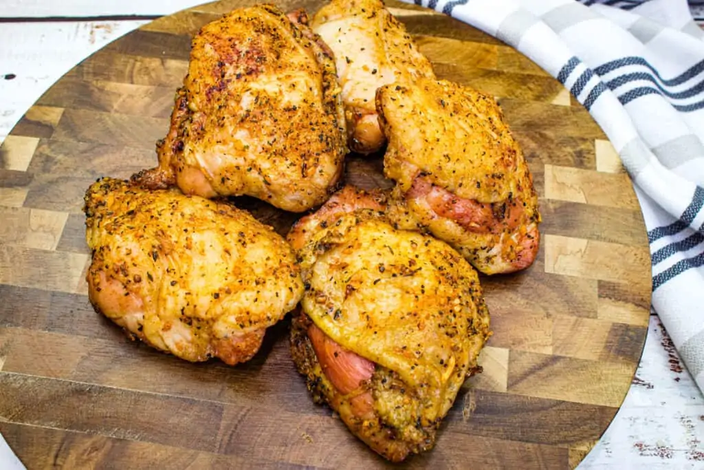Five pieces of seasoned, smoked chicken thighs rest on a round wooden cutting board next to a white and black striped cloth.