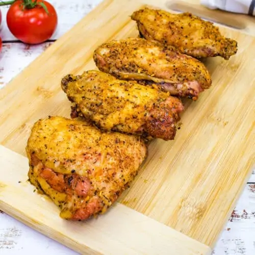 Four smoked chicken thighs, perfectly seasoned and cooked, rest on a wooden cutting board alongside vine tomatoes and a striped towel.