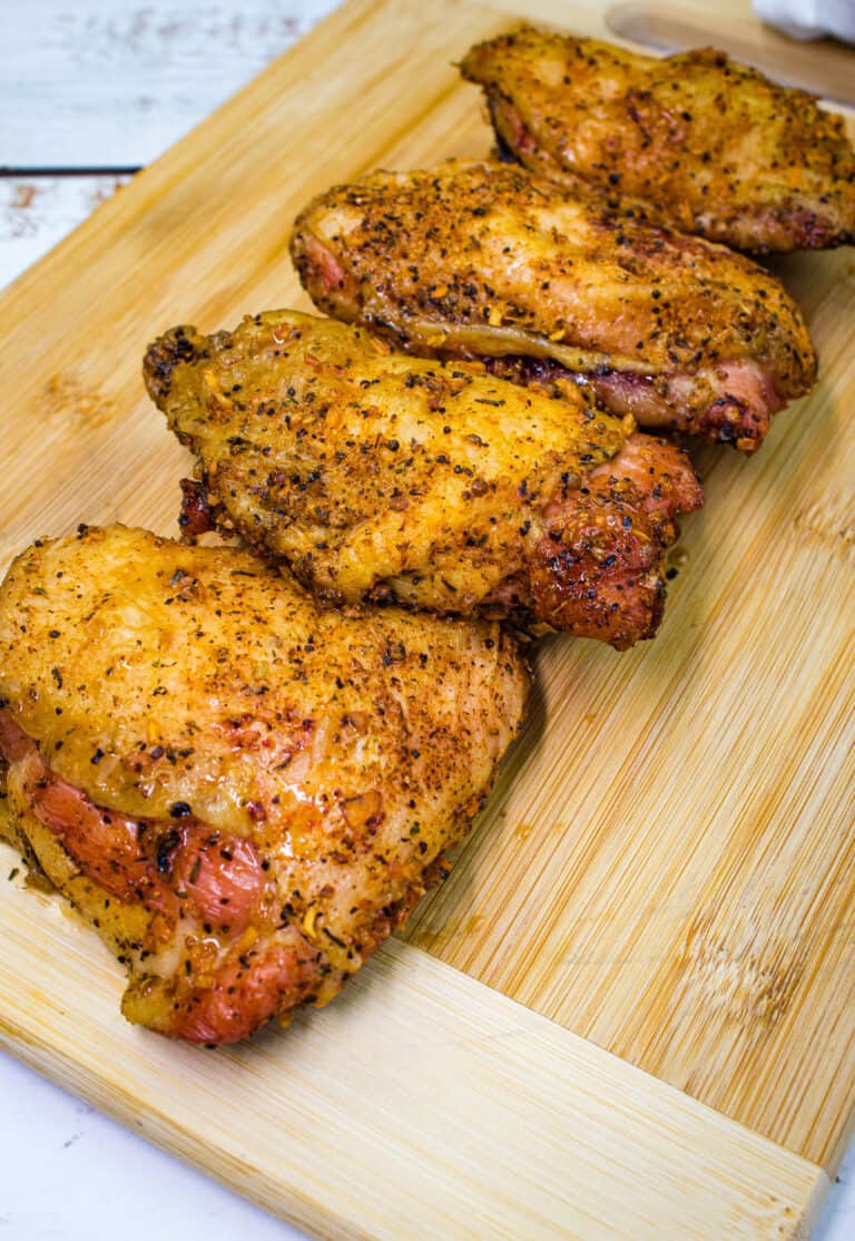 Four seasoned, smoked chicken thighs are arranged in a row on a wooden cutting board.