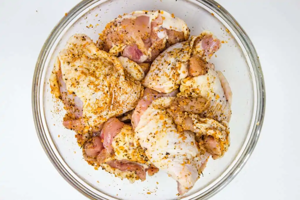 Raw chicken pieces, including smoked chicken thighs, coated with seasoning and spices in a clear glass bowl, photographed from above against a white background.