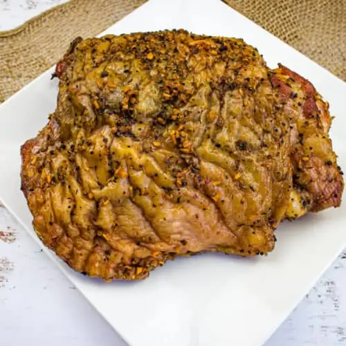 A cooked, seasoned smoked turkey thigh sits on a white square plate atop a piece of burlap on a rustic white wooden table,
