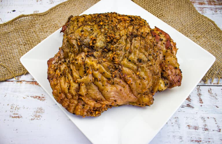A cooked, seasoned smoked turkey thigh sits on a white square plate atop a piece of burlap on a rustic white wooden table,