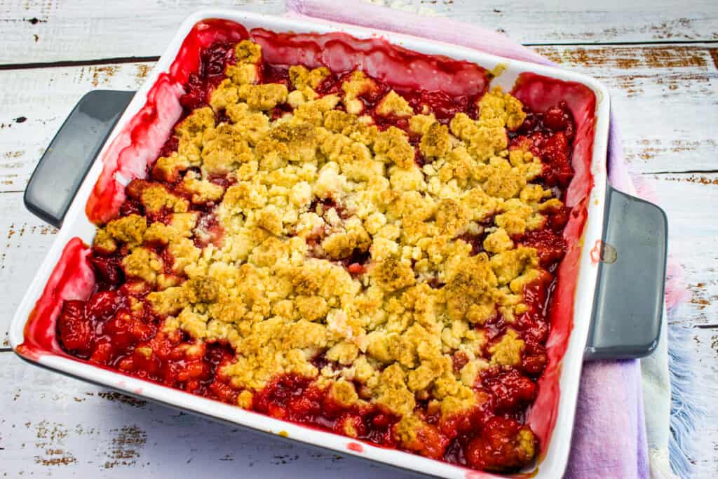 A baked rhubarb and strawberry crumble dessert in a square white dish with golden-brown topping and vibrant red fruit filling visible around the edges.