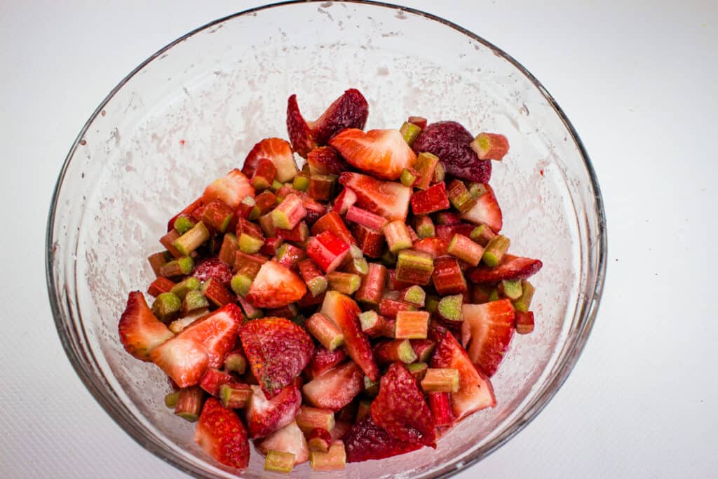 A glass bowl filled with chopped fresh strawberries and rhubarb pieces, lightly coated with sugar—perfect for making a delicious rhubarb and strawberry crumble.