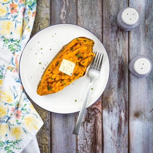 A baked sweet potato, prepared using the bake sweet potatoes in an air fryer method, is topped with butter and herbs on a white plate with a fork, beside a floral napkin and salt and pepper shakers on a wooden table.