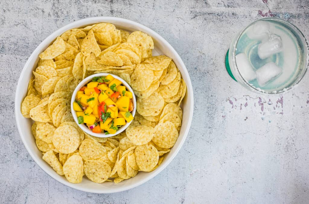 A bowl of tortilla chips surrounds a smaller bowl of mango habanero salsa; a glass of ice water sits nearby on a textured white surface.