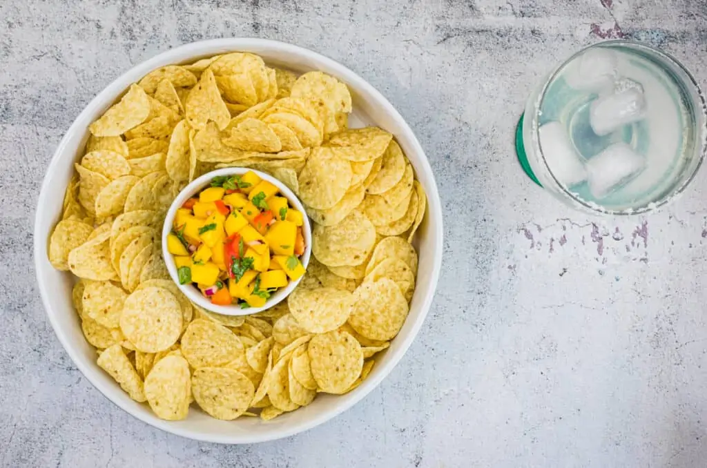 A bowl of tortilla chips surrounds a smaller bowl of mango habanero salsa; a glass of ice water sits nearby on a textured white surface.