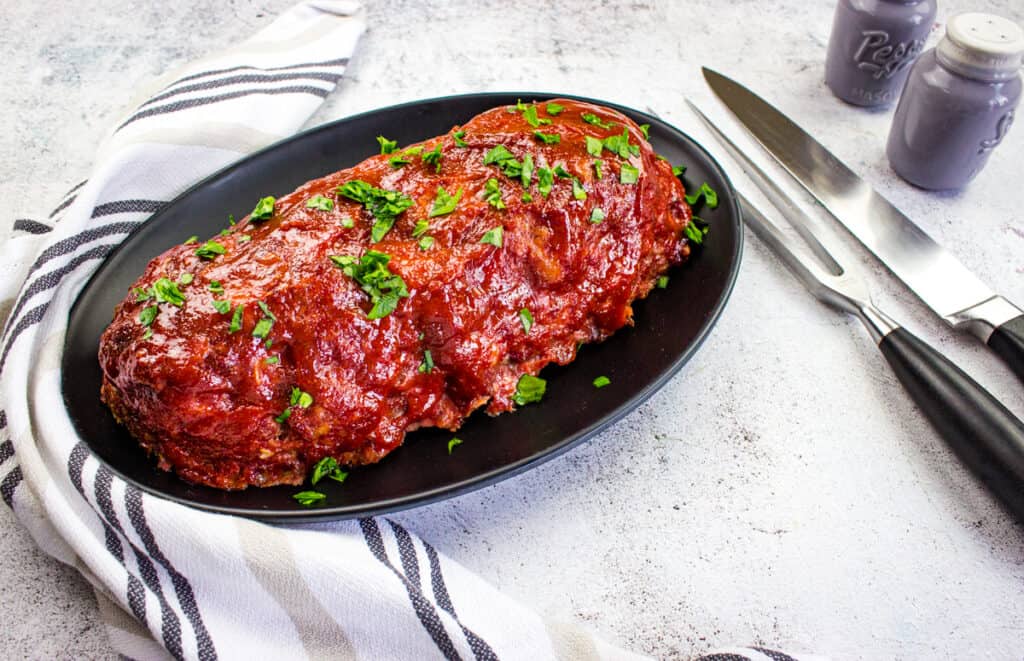 A smoked meatloaf, glazed and topped with chopped parsley, sits on a black oval plate beside a striped towel, carving fork, knife, and salt and pepper shakers.