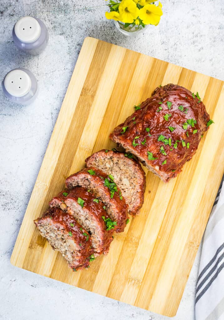 Sliced smoked meatloaf garnished with herbs on a wooden cutting board, alongside salt and pepper shakers and a small vase of yellow flowers.