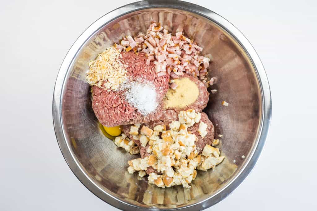 A metal mixing bowl containing ground meat, chopped onions, bacon, breadcrumbs, seasonings, and an egg&mdash;ready to be combined for a delicious smoked meatloaf.