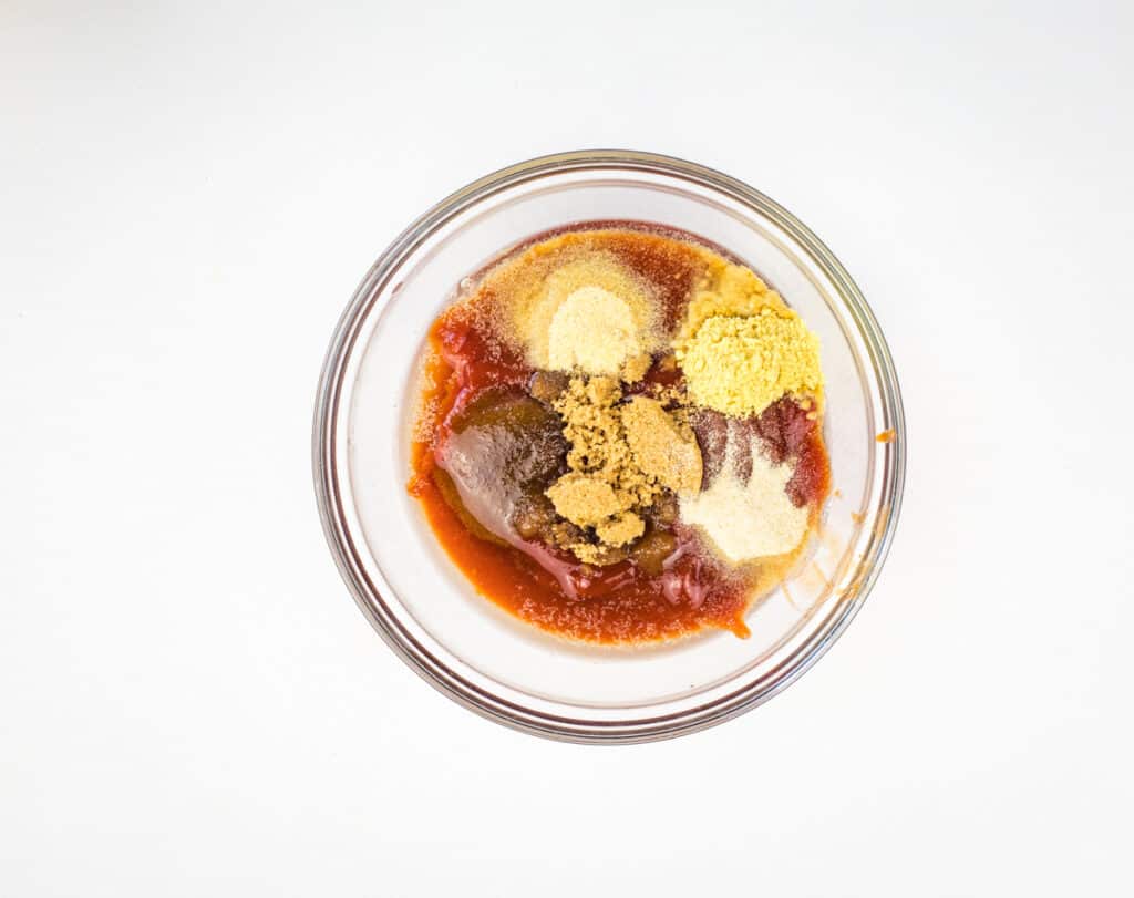 A glass bowl containing ketchup, brown sugar, and various powdered seasonings&mdash;perfect for glazing smoked meatloaf&mdash;set on a white background.