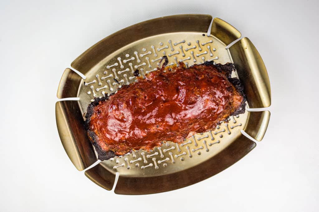 A smoked meatloaf with a glossy glaze sits on a golden roasting tray with a patterned surface, photographed from above on a white background.