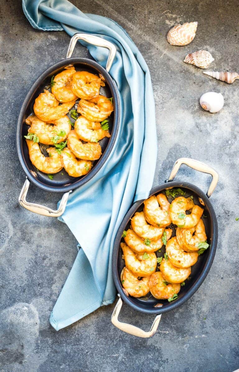 Two oval serving dishes with smoked shrimp garnished with herbs, placed on a gray surface with a blue cloth and decorative seashells nearby.