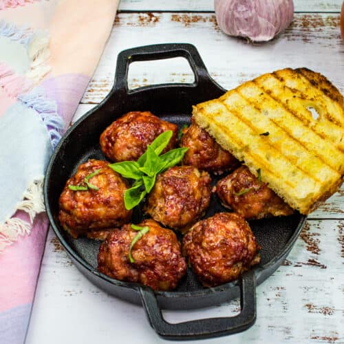 A black skillet with seven smoked meatballs garnished with basil and a slice of grilled bread, placed on a rustic white wooden surface.