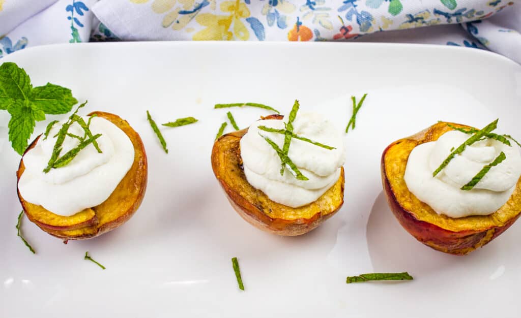 Three roasted peach halves topped with smoked peaches with spiced rum whipped cream and thin mint strips are arranged on a white plate with a mint garnish.