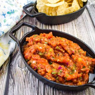 A black dish of chunky smoked salsa sits in front of a matching dish filled with tortilla chips on a wooden surface, next to a floral-patterned cloth.
