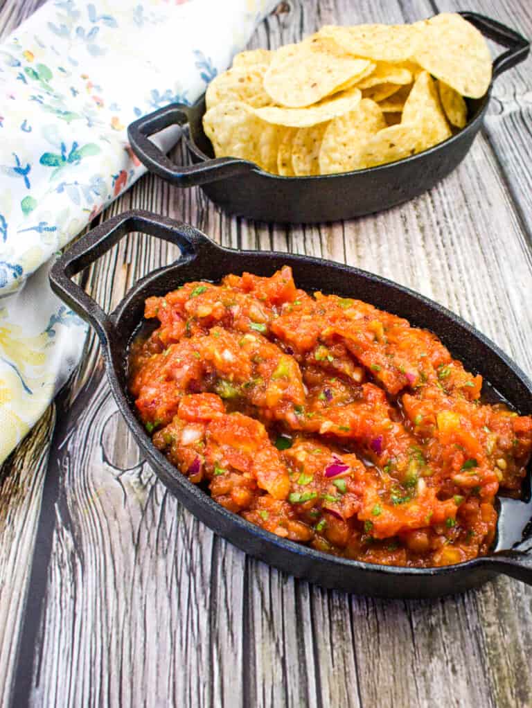 A black dish of chunky smoked salsa sits in front of a matching dish filled with tortilla chips on a wooden surface, next to a floral-patterned cloth.