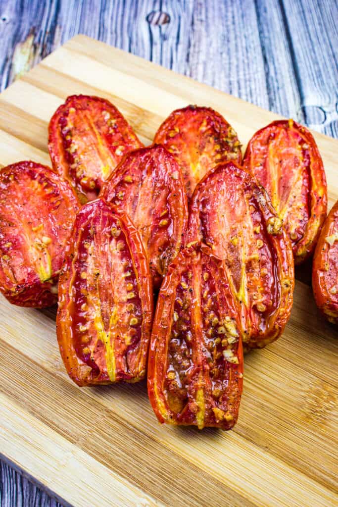 Halved smoked tomatoes with visible seeds are arranged on a wooden cutting board, set atop a table with a wood grain pattern.