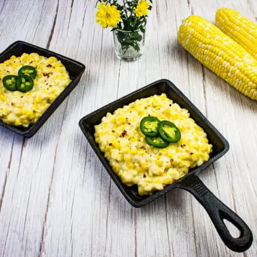 Two cast iron pans filled with creamed corn, topped with sliced jalapeños, sit beside raw corn cobs and a small vase of yellow flowers on a white wooden surface.