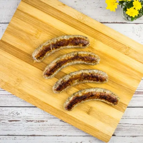 Four sous vide sausages are arranged in a diagonal line on a wooden cutting board, with a small vase of yellow flowers in the top right corner.