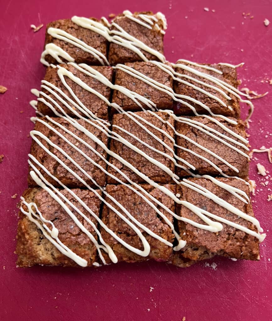 Fifteen chocolate brownies cut into squares, arranged on a red surface, and drizzled with white icing&mdash;perfect alongside cranberry bars for a festive treat.