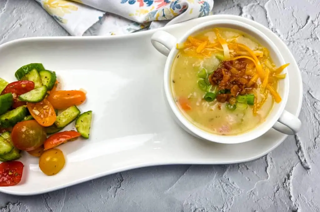 A fully loaded baked potato soup served in a white plate with fresh vegetables.