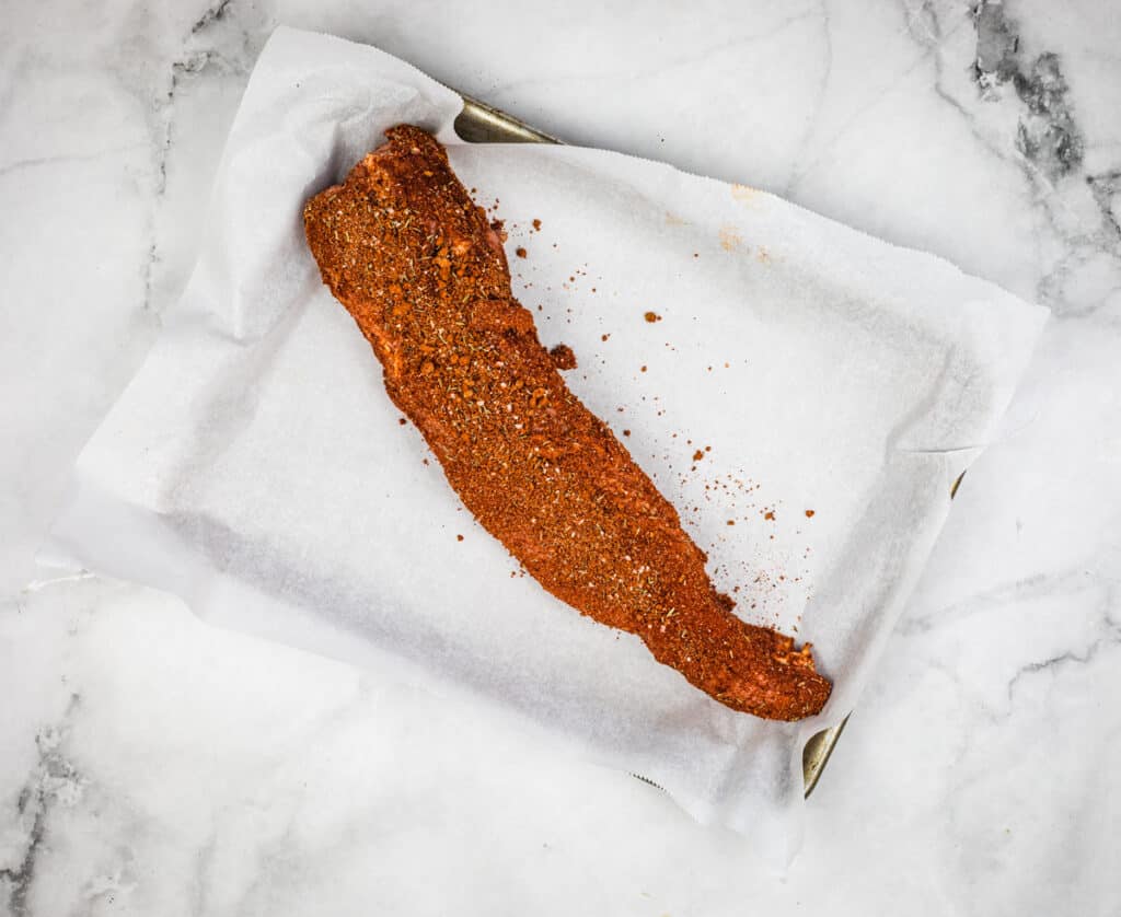 Uncooked pork tenderloin liberally coated with rub on a sheet pan.