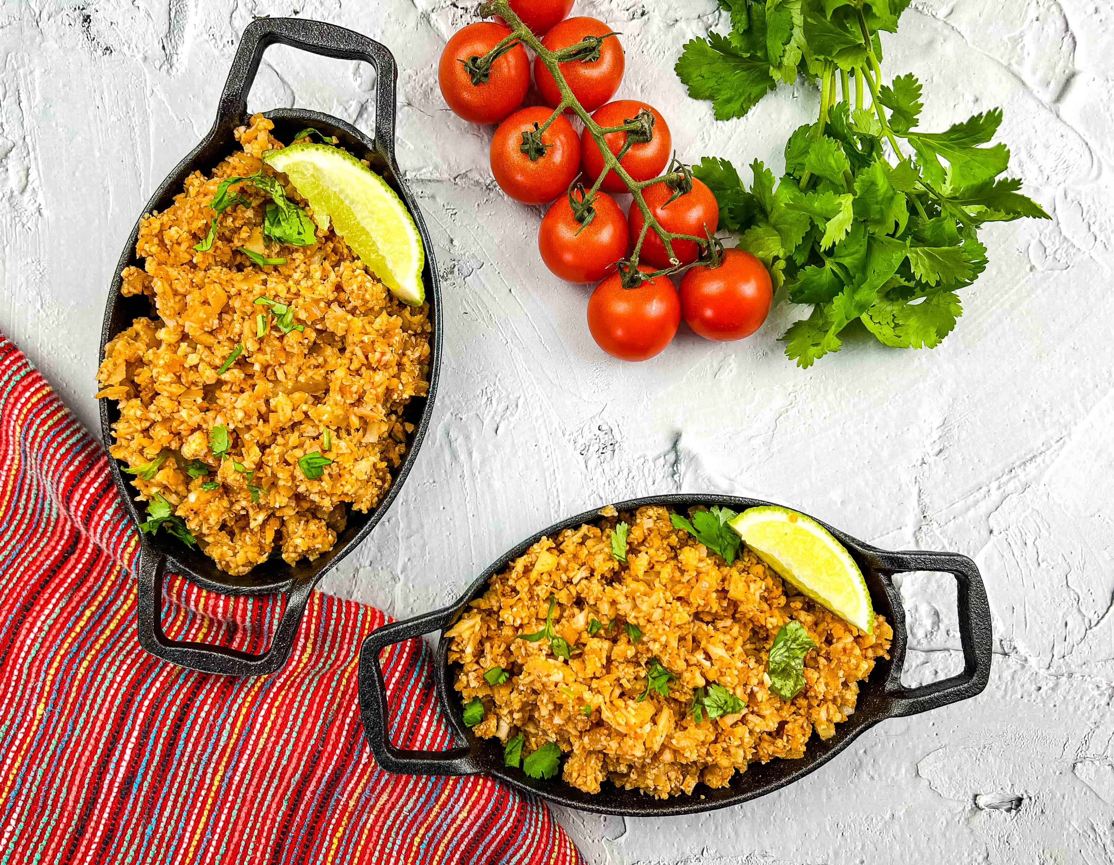 A plate of Mexican Cauliflower Rice garnished with cilantro, served with a lime wedge. Fresh tomatoes and cilantro are visible in the background.