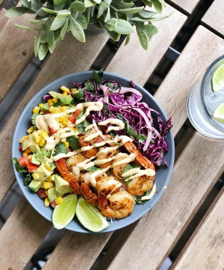 A bowl with grilled shrimp, sliced avocado, corn, tomato, shredded red cabbage, greens, lime wedges, and a drizzle of sauce, on a wooden table next to a glass of water with lime.
