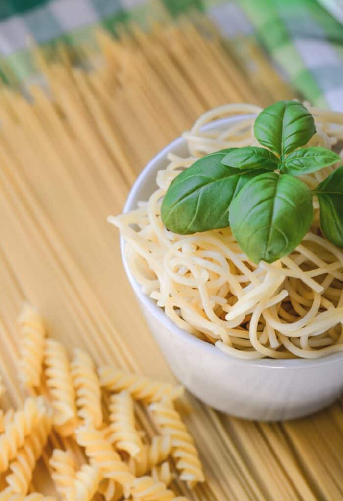 A white bowl of cooked spaghetti topped with fresh basil sits on a surface with uncooked spaghetti and rotini pasta in the background.