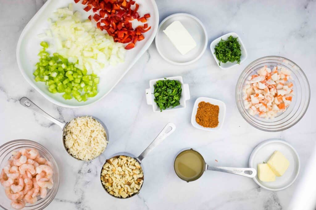 Chopped onions, red bell pepper, celery, shrimp, parsley, rice, breadcrumbs, crab, seasonings, broth, and butter arranged in bowls and measuring cups on a white counter for a flavorful seafood dressing.