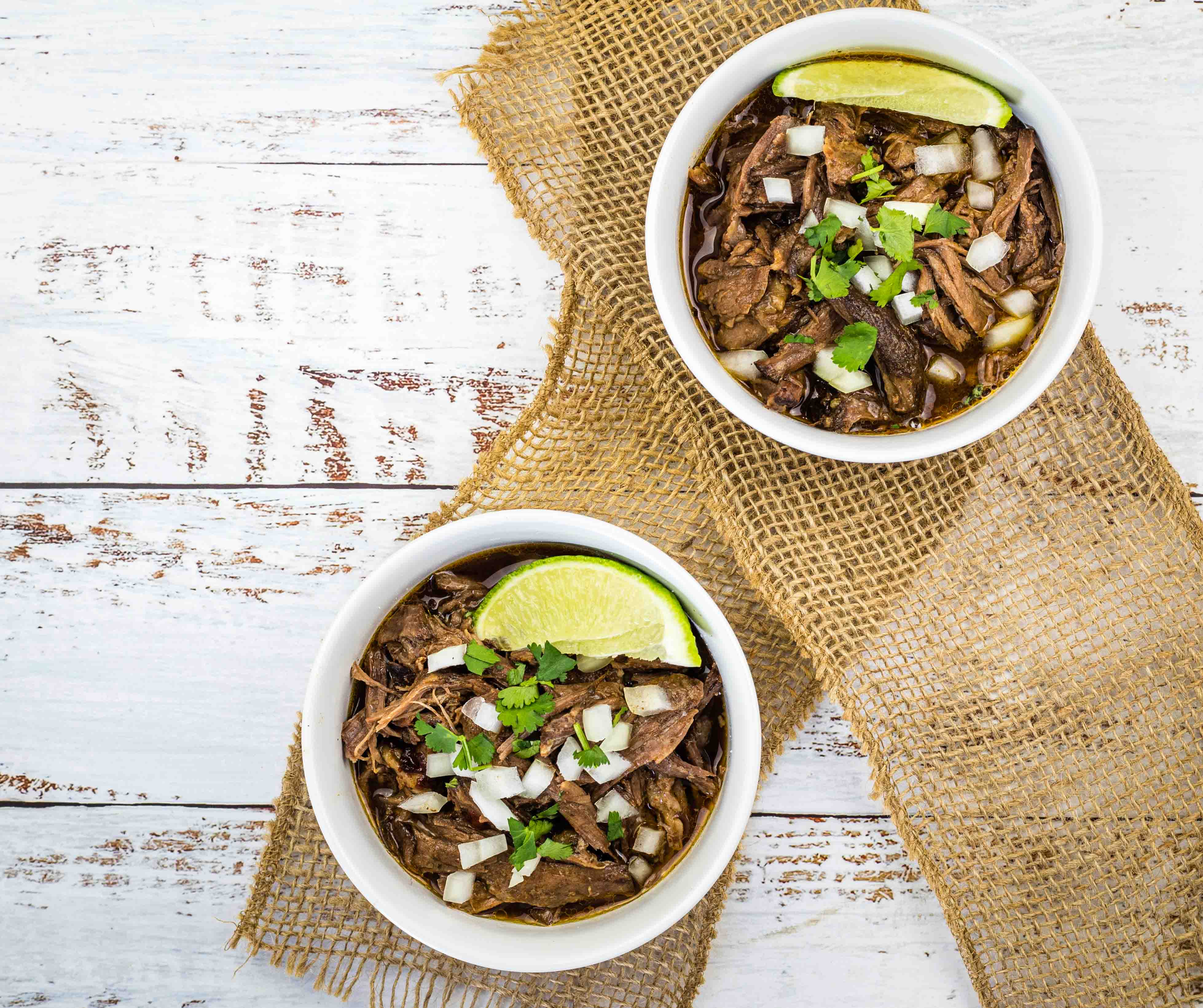 Two white bowls filled with slow cooker birria garnished with fresh lime wedges.
