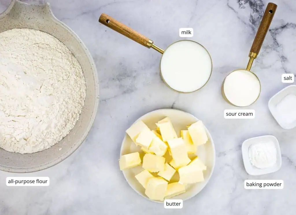 A bowl of flour, a plate of cubed butter, a measuring cup of milk, a smaller cup of sour cream, and small bowls of salt and baking powder are arranged on a marble surface, ready to craft delicious smoked biscuits.