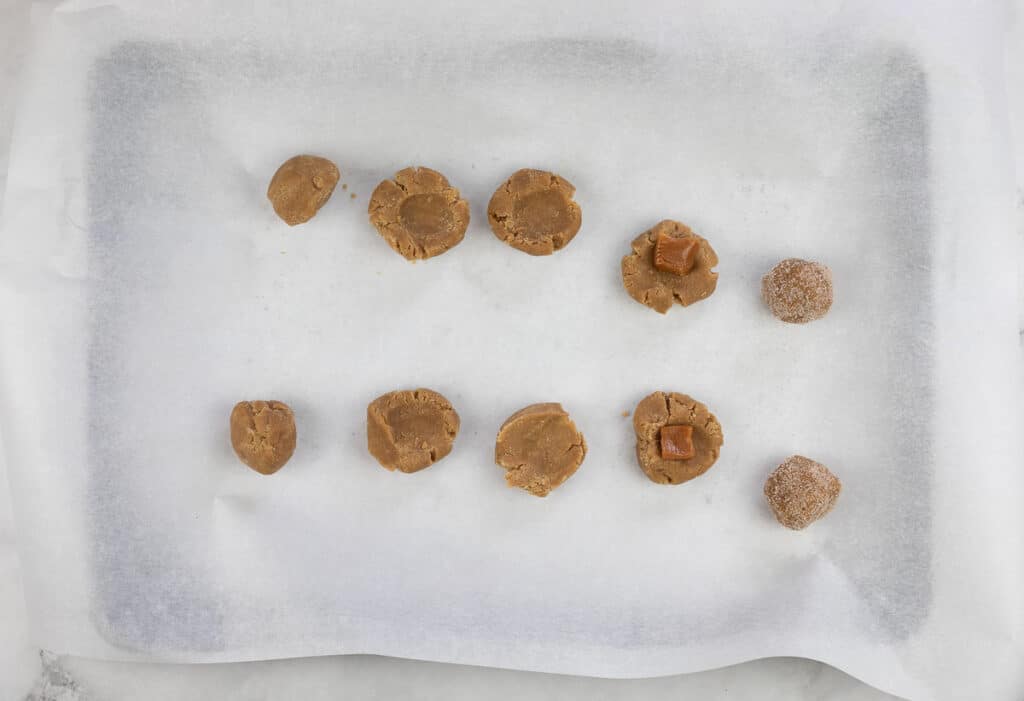 A baking sheet lined with parchment paper holds nine unbaked salted caramel brown butter snickerdoodle dough balls, some rolled in sugar and some studded with caramel pieces.