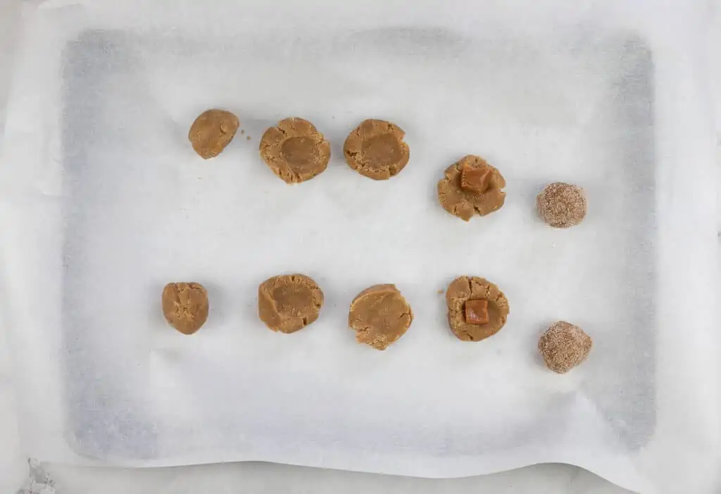 A baking sheet lined with parchment paper holds nine unbaked salted caramel brown butter snickerdoodle dough balls, some rolled in sugar and some studded with caramel pieces.