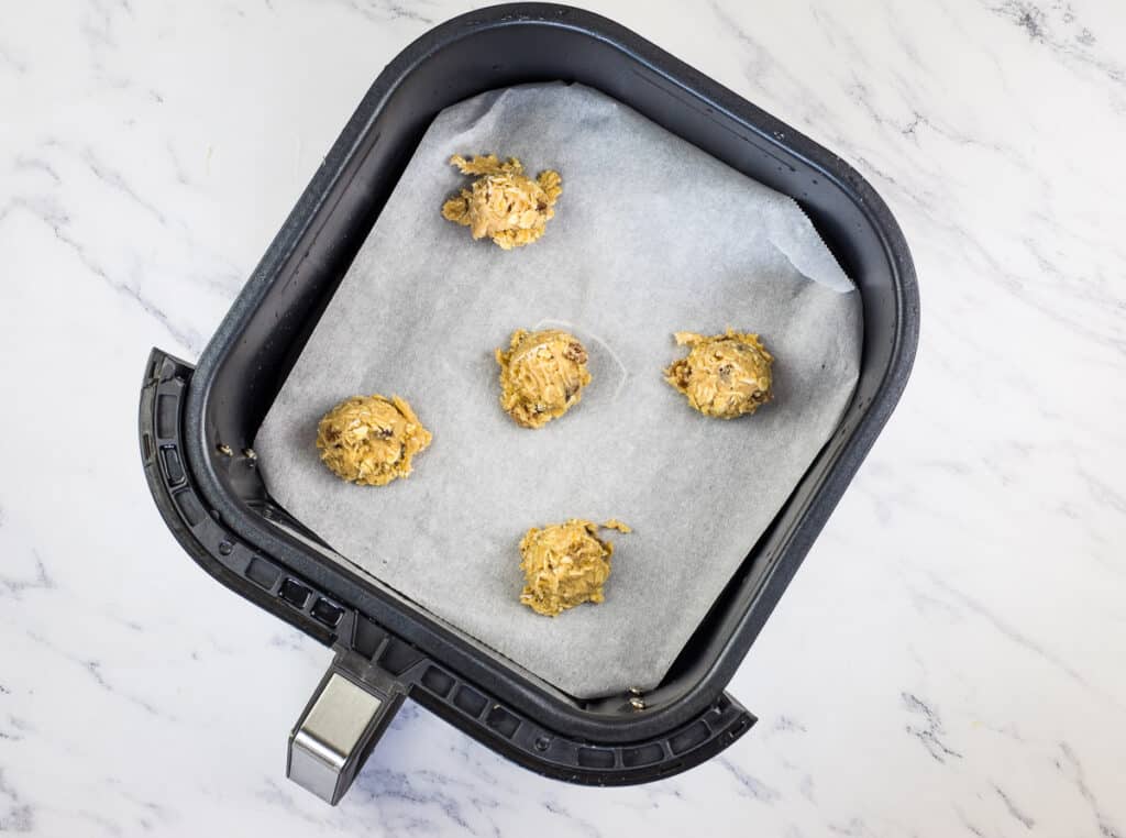 Five scoops of raw air fryer oatmeal raisin cookie dough placed on parchment paper inside an air fryer basket, ready to be baked.