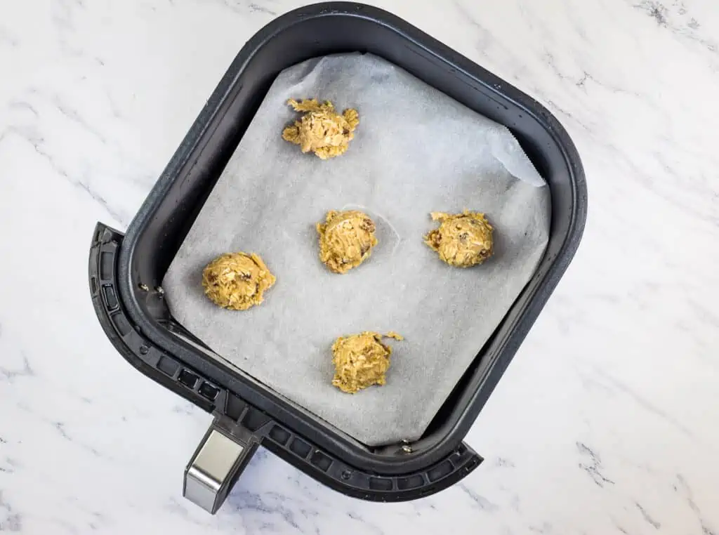 Five scoops of raw air fryer oatmeal raisin cookie dough placed on parchment paper inside an air fryer basket, ready to be baked.
