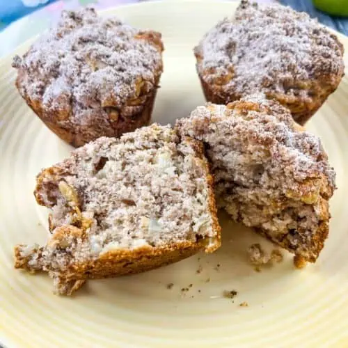 A sliced Apple, Date and Walnut muffin on a plate with 2 muffins in the background.