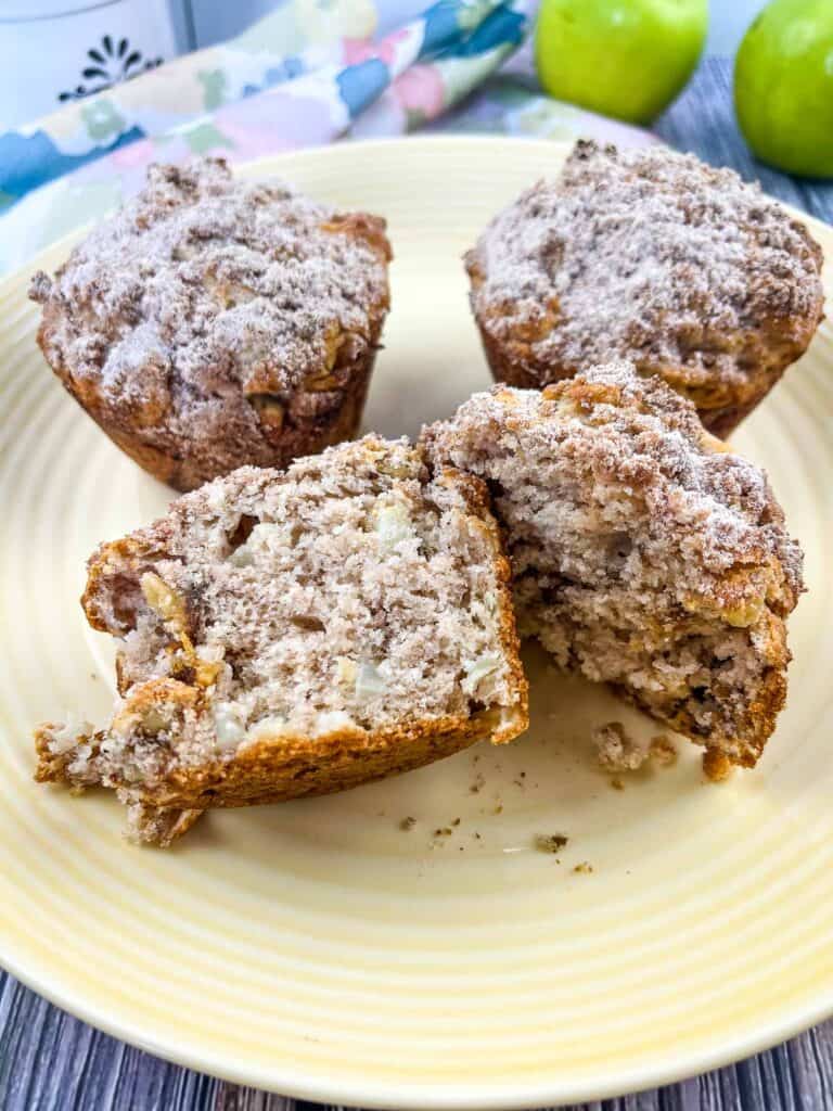 A sliced Apple, Date and Walnut muffin on a plate with 2 muffins in the background.