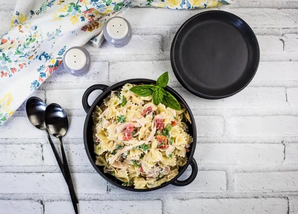 A top-down shot of chicken pasta salad in a black serving bowl with serving plates to the side.