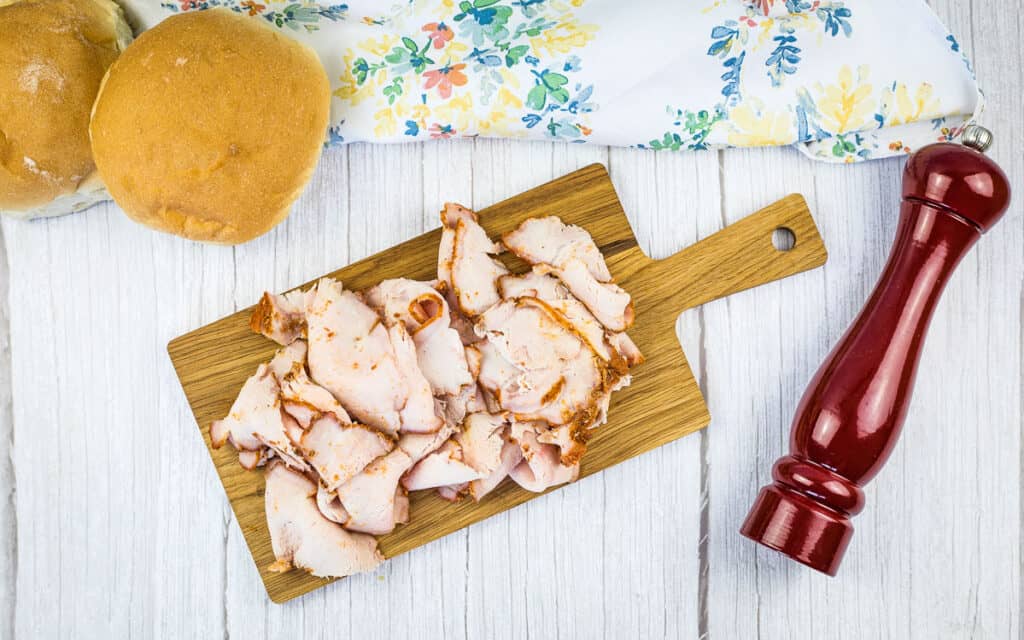 A top down shot of smoked pork lunch meat on a cutting board with buns in the background.